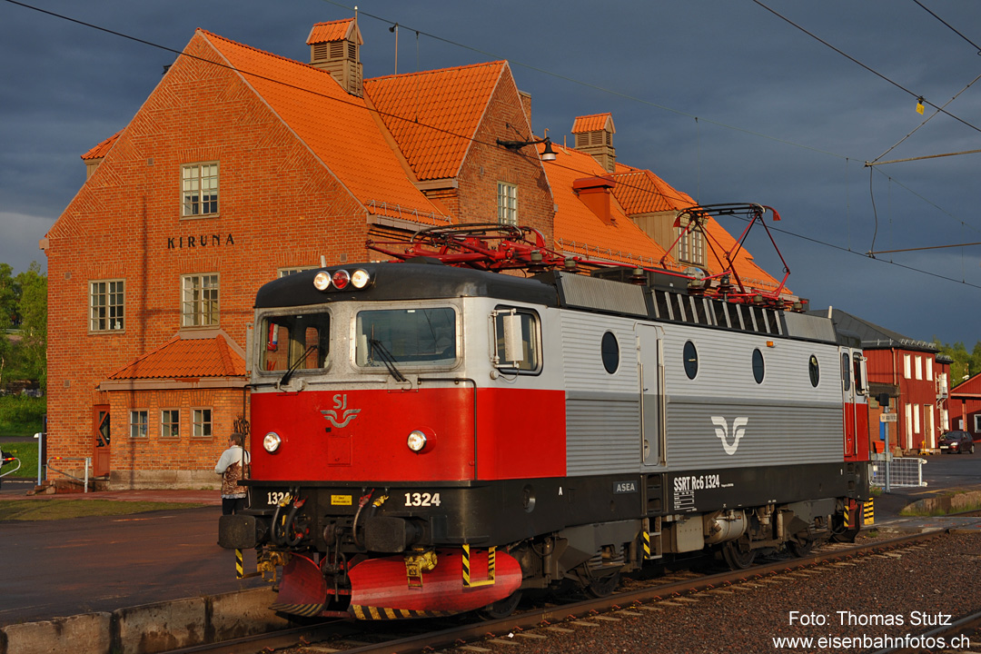 Lok mit Bahnhofsgebäude
Während des Umfahrens leuchtet das Bahnhofsgebäude (durch die Sonneneinstrahlung) vor den dunklen Regenwolken.
